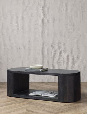 The Luna black wood oval coffee table sits in front of a textured white wall on chevron hardwood flooring with an open book sitting at the base of the table with a book and white bowl sitting on its surface.