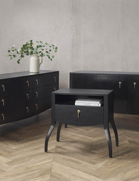 | The Anabella black wood nightstand sits in a studio with books sitting in its open shelf and is next to the Anabella dresser