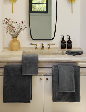 The Midnight gray dune turkish cotton Air Weight Towel Set by Coyuchi hangs off of a concrete sink in a bathroom with a narrow black framed mirror and a sculptural yellow vase