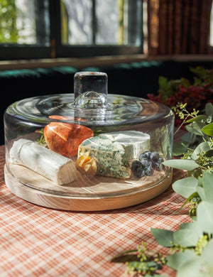 The Lotta cheese and pastries glass dome with ash wood base by LSA International sits in a dining room atop a red plaid tablecloth next to an olive garland