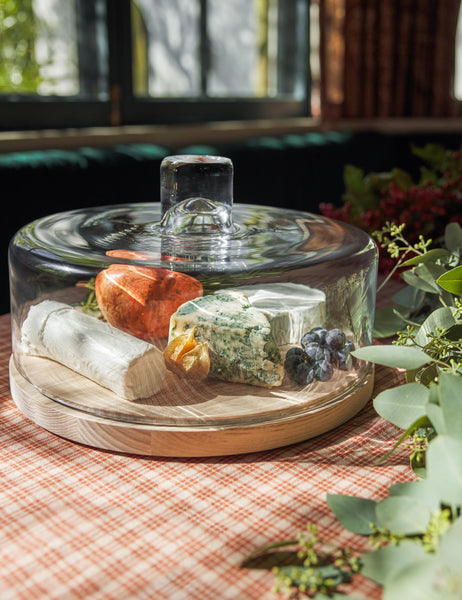 | The Lotta cheese and pastries glass dome with ash wood base by LSA International sits in a dining room atop a red plaid tablecloth next to an olive garland