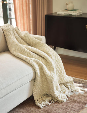 The Olema ivory handwoven throw with fringed ends lays in a living room atop a natural linen sofa and a brown area rug with a black sideboard in the background