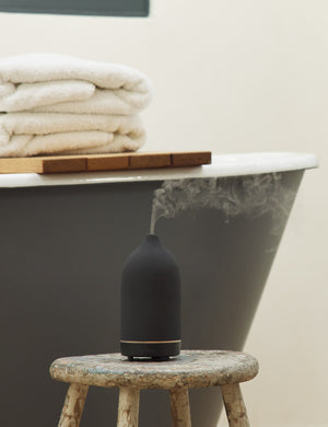 The Black Stone Diffuser by Vitruvi sits atop a wooden stool in a bathroom with a black painted tub