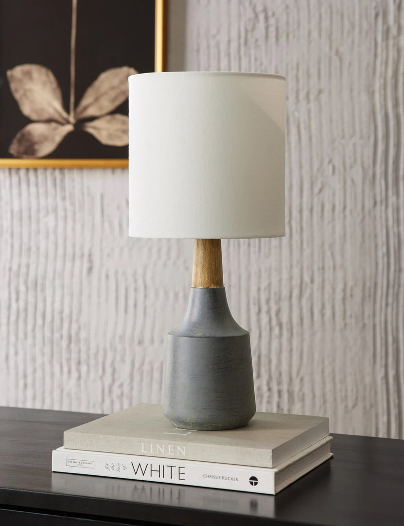 #color::stone | The Marcella gray vase-shaped Mini Table Lamp with ceramic base sits on top of a wooden sideboard with a stack of books and a floral wall art in the background