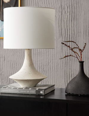 The Coulwood ivory table lamp with sculptural base sits on a black sideboard atop a gray book with a sculptural black vase