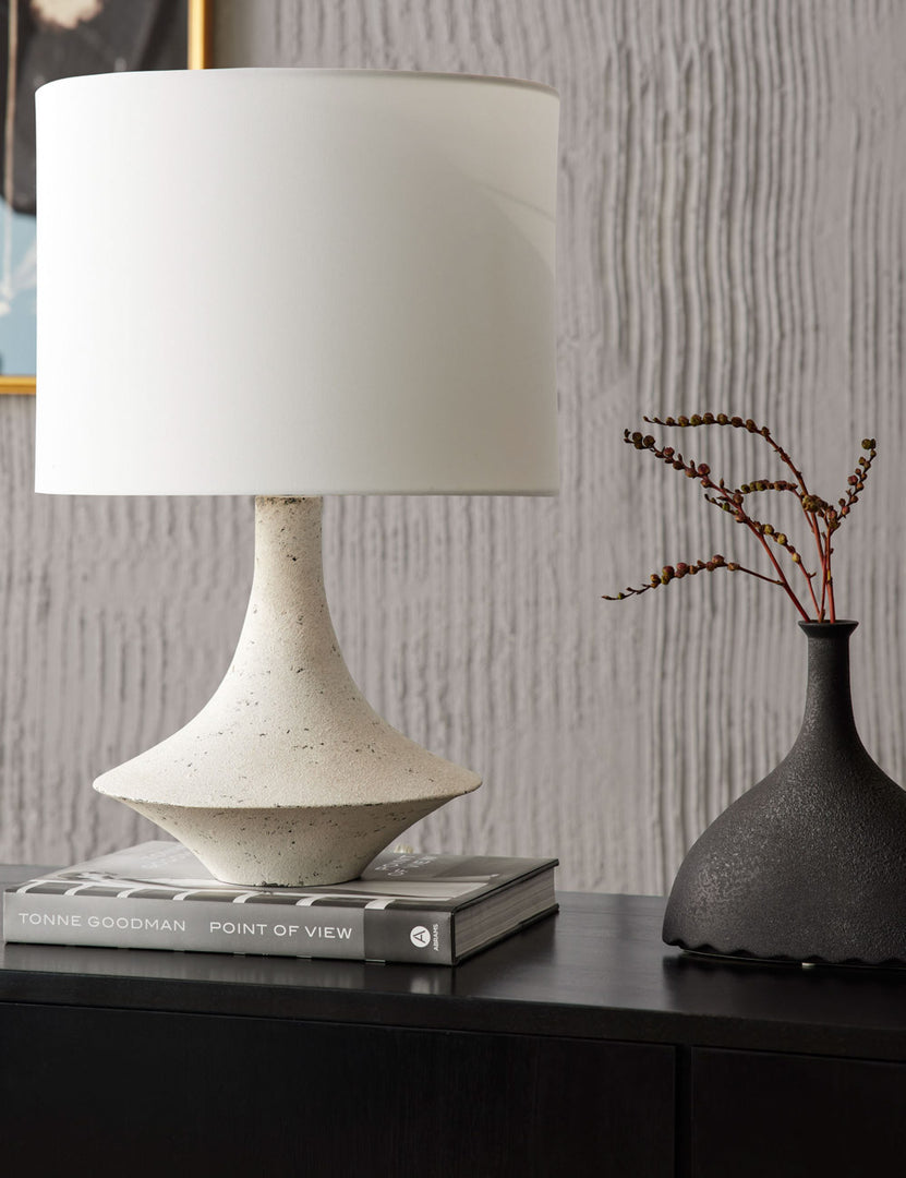 #color::ivory | The Coulwood ivory table lamp with sculptural base sits on a black sideboard atop a gray book with a sculptural black vase