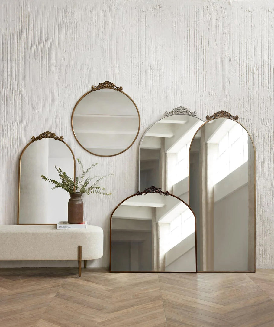 #silver | The Tulca silver curved standing mirror with flat bottom edge and traditional scroll detailing sits on a chevron hardwood floor surrounded by other Tulca mirrors in their circular, narrow, and standard sizes. 