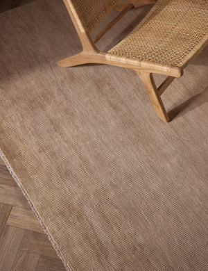 The Heritage wheat rug lays on a chevron wooden floor under a woven accent chair