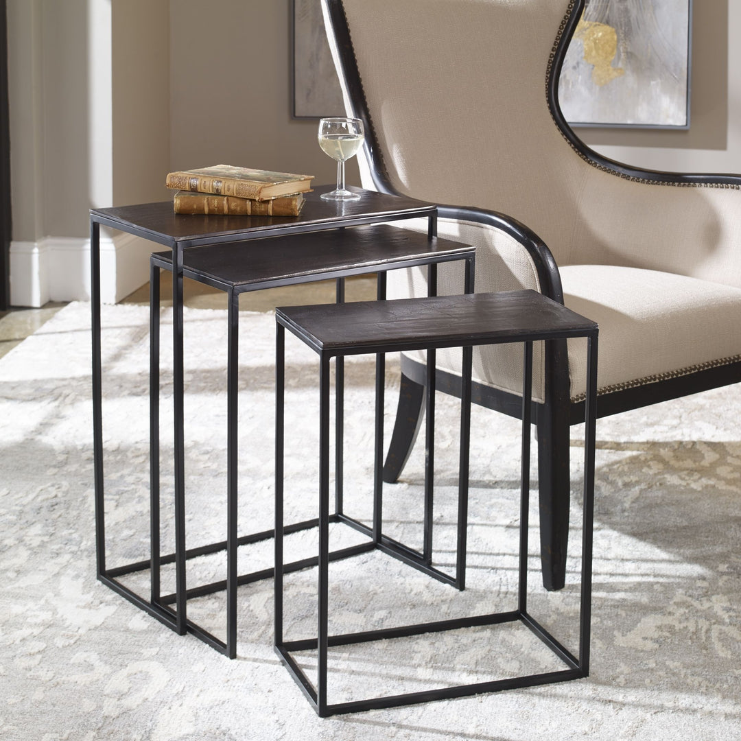 | The Loletta black nesting tables are partially nested under each other, sitting in a living room next to a black wooden framed accent chair atop a gray patterned rug