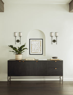 The Lilies Wall Art hangs in between two double sconces above a black sideboard with books and a planter