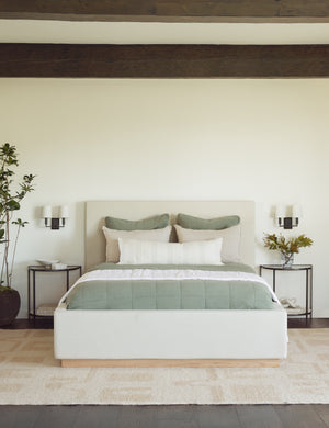 The Lockwood natural velvet-upholstered bed with a white oak base sits in a neutral bedroom with a plush natural rug and wooden ceiling beams.