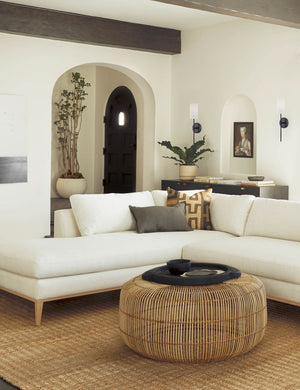 The Jensine old bronze slim sconce with cylindrical shade is mounted against a white wall in a living room with a white sectional, a woven coffee table, and a natural jute rug