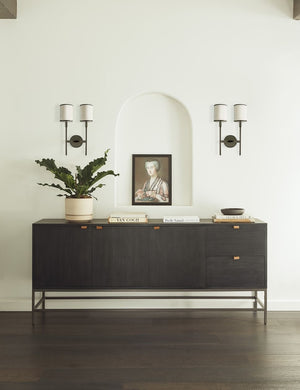 The Rosamonde black wood sideboard with brown leather pulls and a metal base sits under an arched cut-out with a painting of a woman inside, flanked by two torchiere wall sconces.