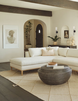 The Sara singh back drop portrait neutral toned wall art by Stampa is hung in a living room with a white linen sectional sofa, a round wooden coffee table, and a textured beige rug