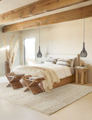The Uma taupe and natural checkered grid pattern rug lays in a bedroom with wooden-beamed ceilings and a linen and wooden framed bed