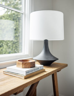 The Coulwood black table lamp with sculptural base sits atop a wooden sideboard with a wooden octagonal bowl and a stack of books