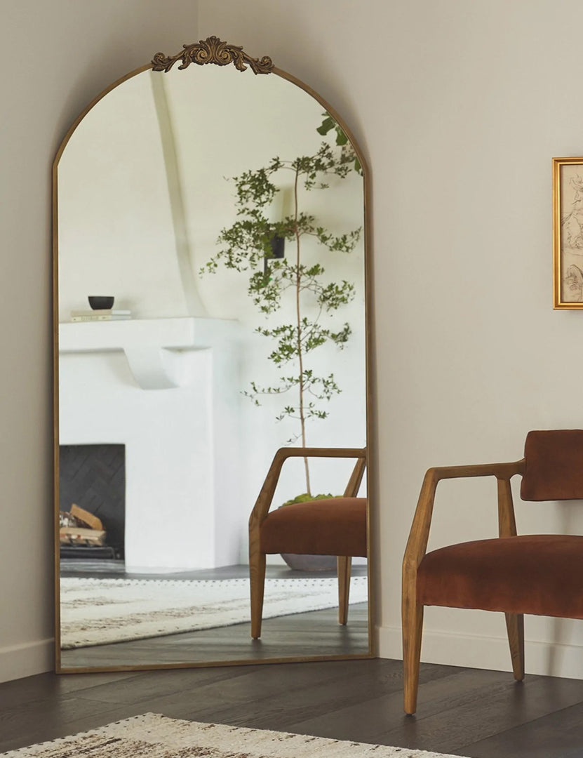 #color::gold | The Tulca gold curved standing mirror with flat bottom edge and traditional scroll detailing sits in the corner of a room on a black hardwood floor with a brown velvet accent chair to the right of it.