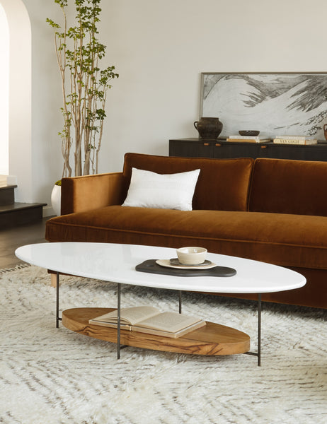 | The Thomas Bina oval coffee table with white laquered top, oak shelf and steel frame sits in front of a dark rust velvet sofa and on top of a neutral area rug.