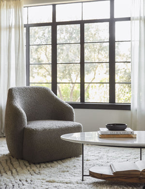 The Tobi gray boucle swivel chair sits atop a plush ivory rug in front of a wall with large black-framed windows and sheet curtains