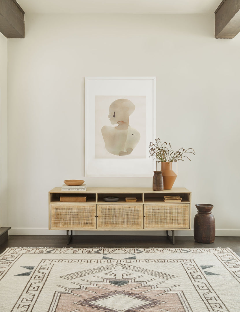#color::natural | The Hannah natural wood media console with cane doors sits in an open space with a large portrait wall art mounted above it and a geometric woven rug below it.