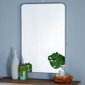 The Lyta rectangular silver framed wall mirror with rounded corners hangs on a blue painted wall above a wooden shelf