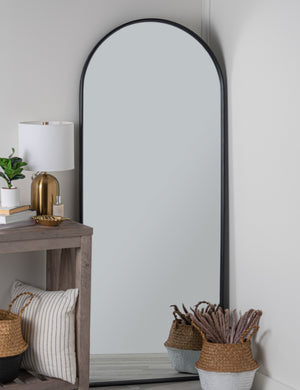 The Shashenka black arched floor mirror sits in the corner of a room with a gold lamp, woven baskets, and a wooden side table