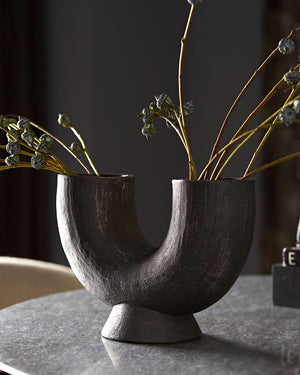 The Damien half-circle sculpture vase by Ateriors with matte black glaze finish sits on top of a stone dining table and is filled with branches