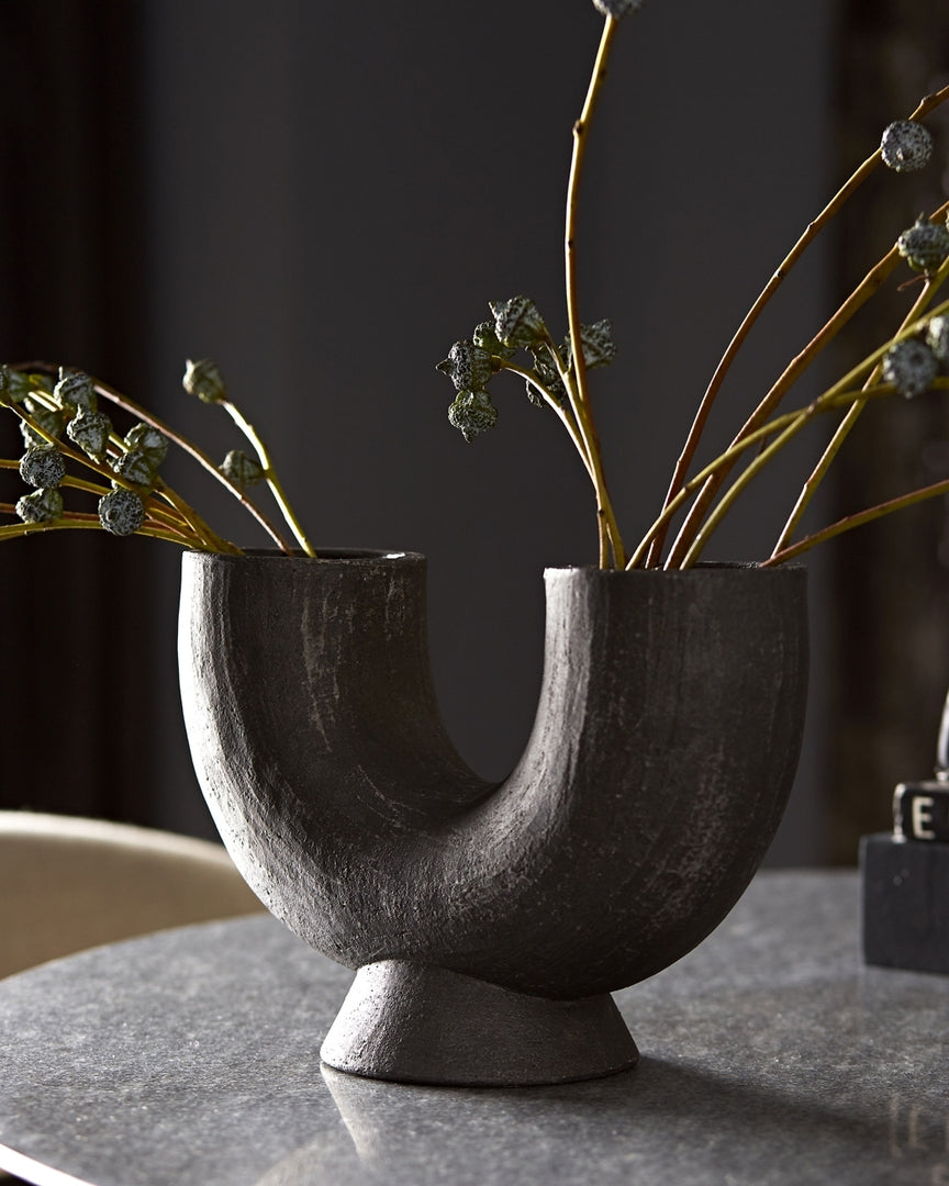 | The Damien half-circle sculpture vase by Ateriors with matte black glaze finish sits on top of a stone dining table and is filled with branches