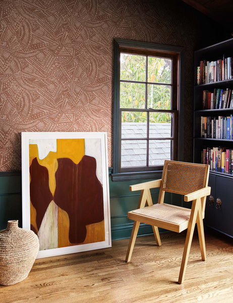 | The Alaari Wallpaper is in a room with a black bookshelf, a woven chair, a jute basket, and a warm-toned wall art