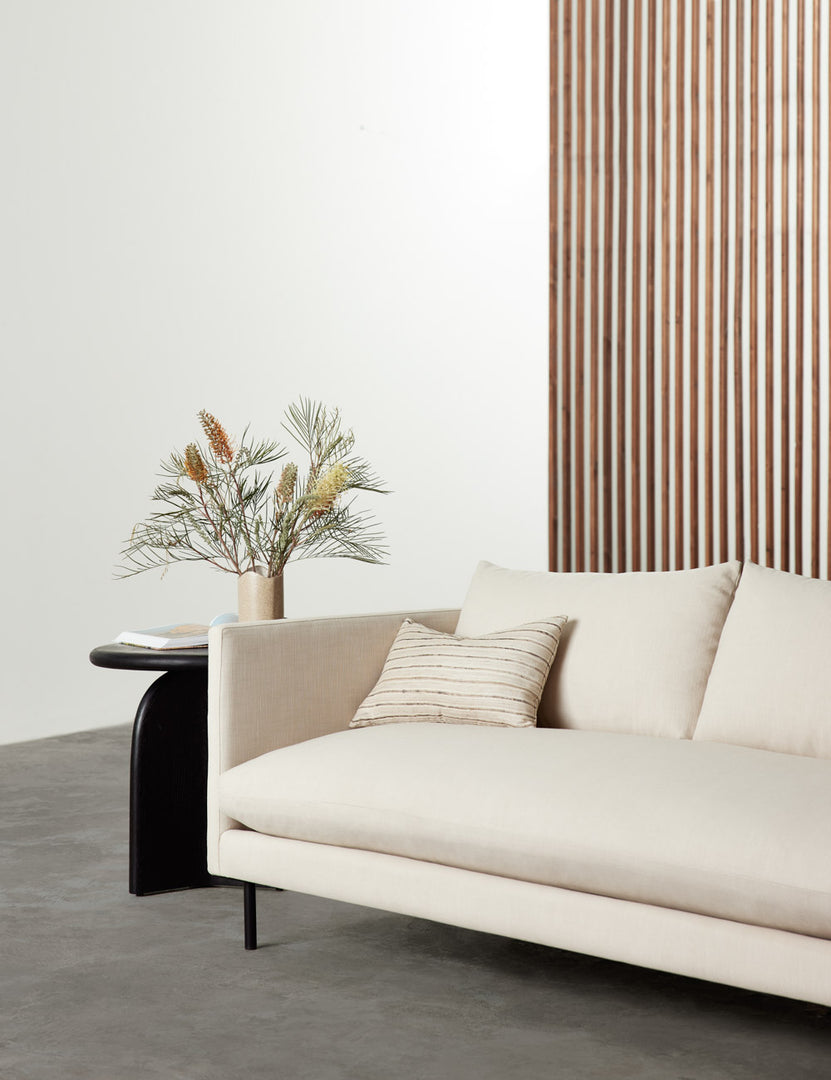 #color::natural #size::72-W #size::84-W #size::96-W | Natural Alaya Sofa sits in a room with a wooden accent wall, a striped throw pillow, and a black side table with a plant