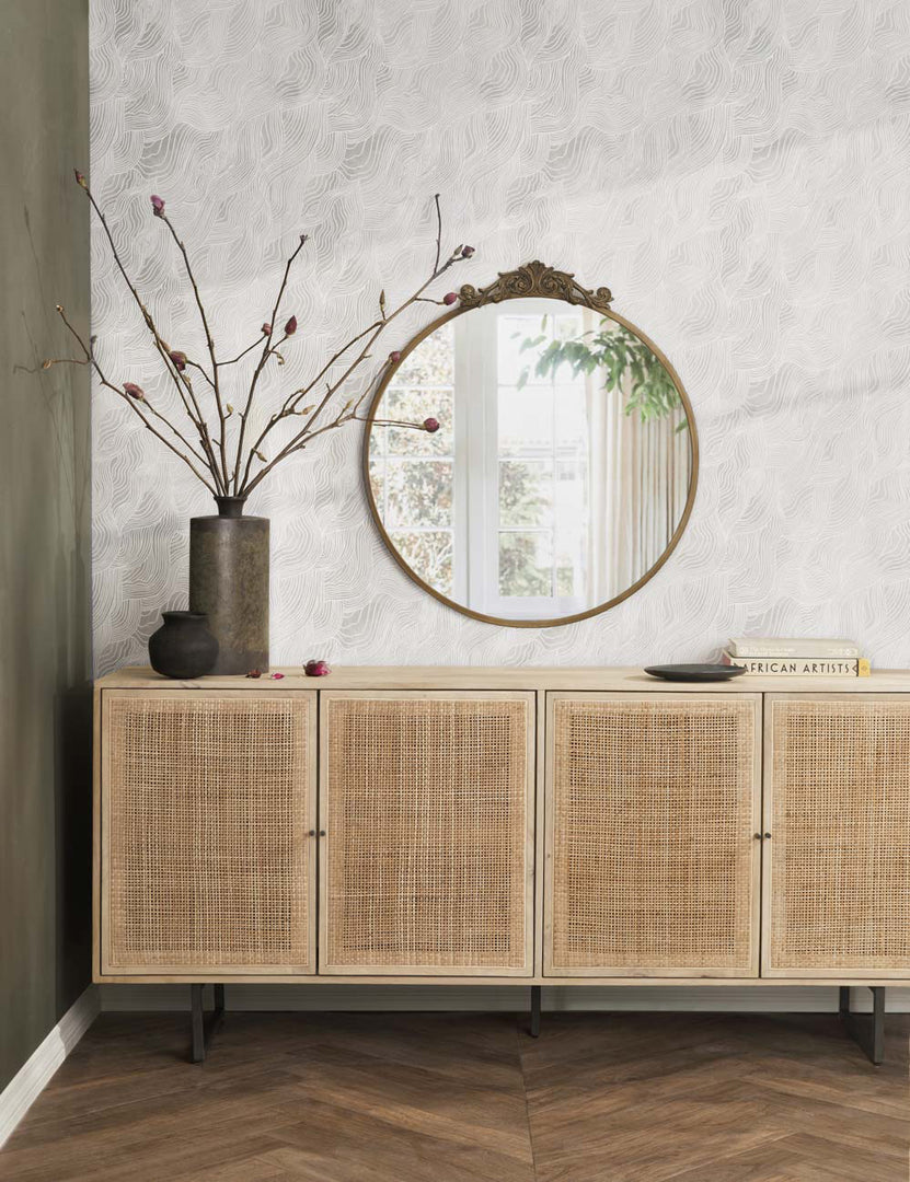 #color::gold | The tulca gold round mirror hands on a wall above a rattan sideboard next to a brown vase