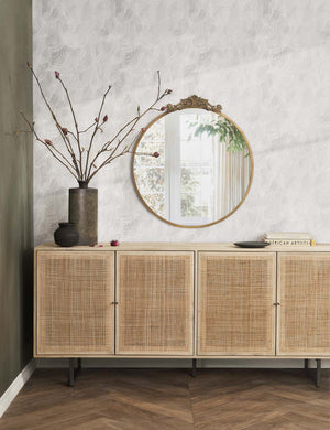 The Hannah natural mango wood sideboard with cane doors sits underneath a circular bronze decorative mirror with a large copper vase sitting atop it on a chevron hardwood floor.