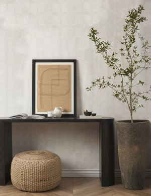 The Vela beige all-natural jute and cotton textural pouf sits in a room under a black wooden console table with a earthenware vase and a brown patterned wall art