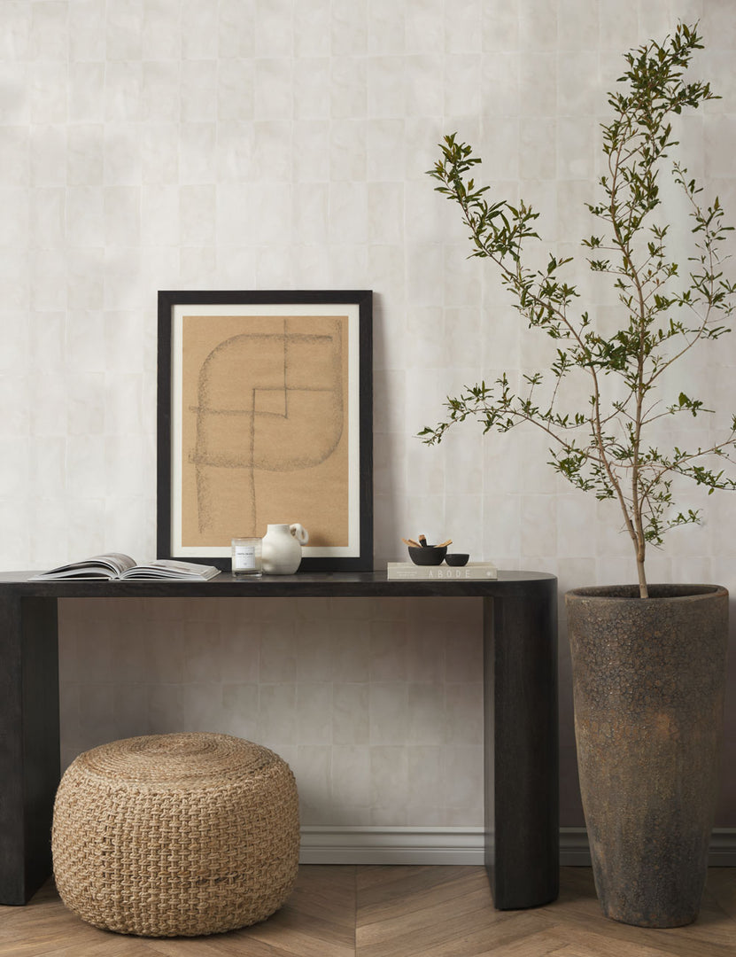 #color::black | The Luna black mango wood oval console table sits in an entryway against an ivory wall with jute woven pouf underneath and a tall ceramic vase to the right.
