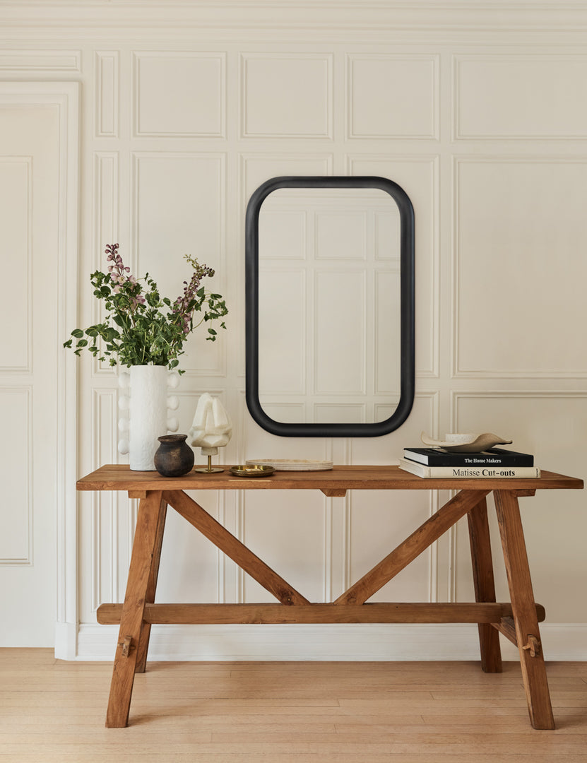 | The Arlene craftsman-style antiqued teak wood console table sits against a white paneled wall with a black framed mirror above it and a white vase with stems and a stack of books on it.