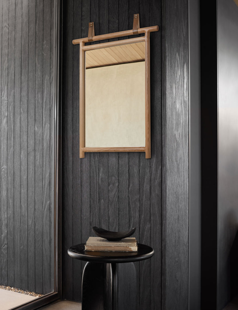 | The kendyl leather strap wall mirror hangs from a black wood paneled wall above a round black side table