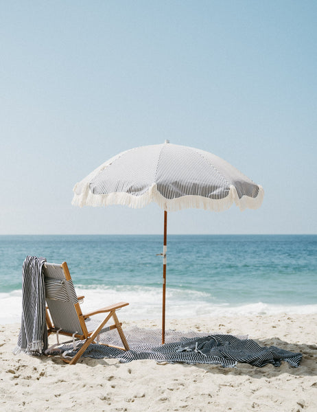 #color::navy-stripe #style::navy-stripe | Navy and white striped Beach Towel by Business & Pleasure Co hangs from a beach chair on a beach with a fringed umbrella