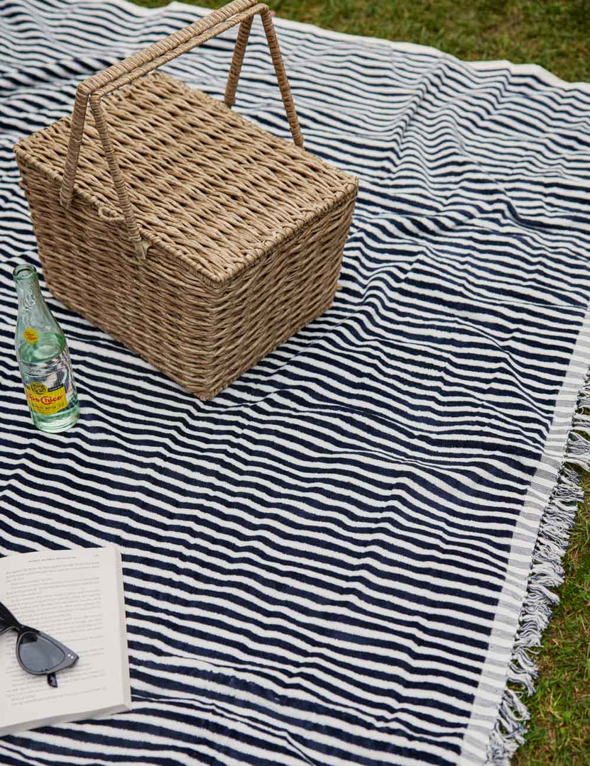 #color::navy-stripe | The Navy and white striped cotton beach blanket by business and pleasure co lays on the grass with a woven picnic basket