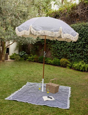 The Navy and white striped cotton beach blanket by business and pleasure co lays on the grass with a striped umbrella standing atop it
