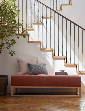 The Grasmere terracotta linen wooden bench sits in front of a staircase with throw pillows, a bowl, and stack of books