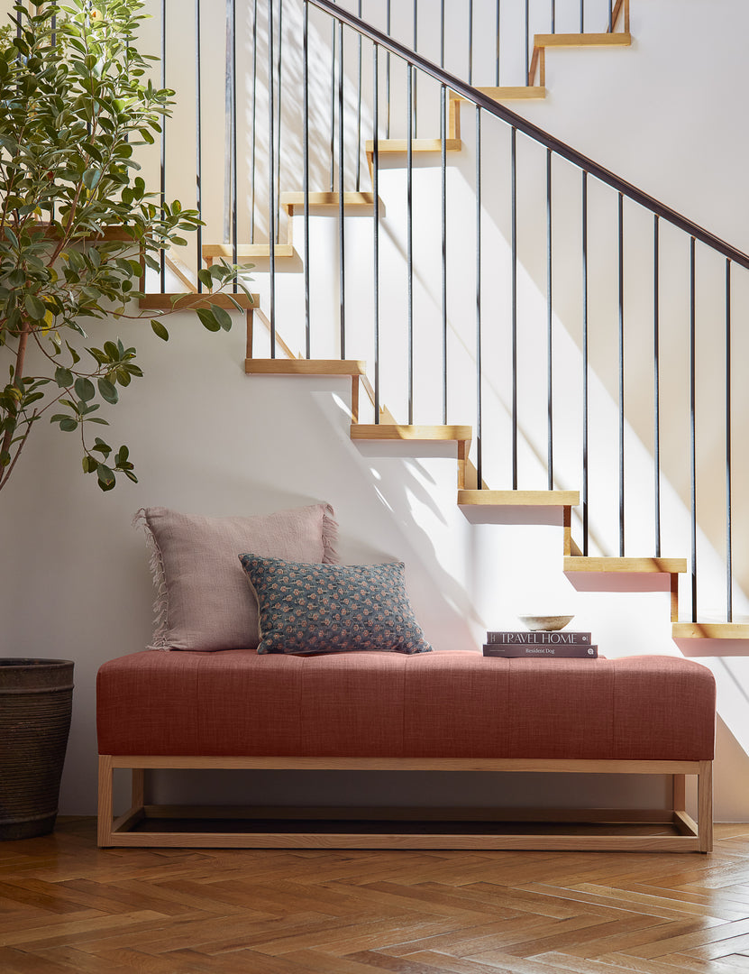 #color::terracotta-linen | The Grasmere terracotta linen wooden bench sits in front of a staircase with throw pillows, a bowl, and stack of books 
