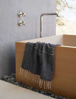 Melkam onyx gray Hand Towel with fringed ends by Bolé Road Textiles hangs off the side of a rectangular wooden tub