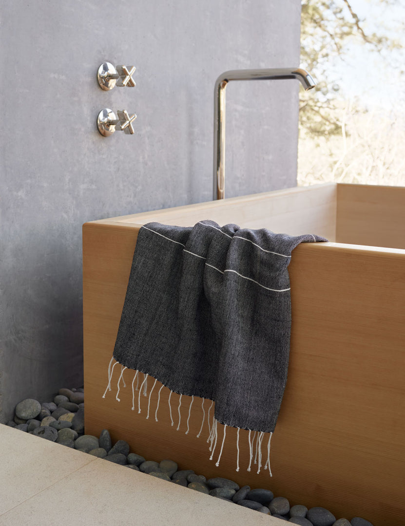 #color::onyx | Melkam onyx gray Hand Towel with fringed ends by Bolé Road Textiles hangs off the side of a rectangular wooden tub