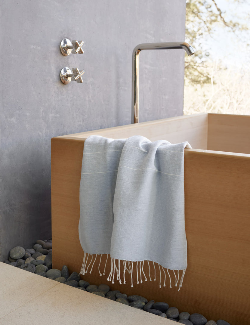 #color::light-gray | Melkam light gray Hand Towel with fringed ends by Bolé Road Textiles hangs off the side of a rectangular wooden tub