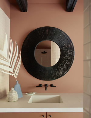 The Carlotta Round Mirror hangs in a bathroom against a terracotta wall above a white sink with black hardware