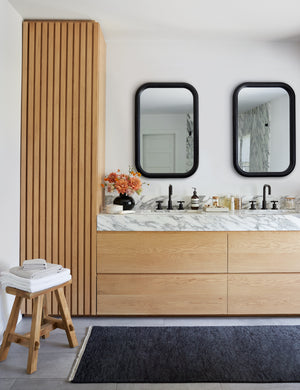 Two bourdon black mirrors hang in a bathroom above his and hers sinks, a navy rug, and a cushioned stool
