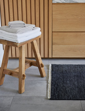 The white turkish cotton Air Weight Towel Set by Coyuchi sits in a stack atop a wooden stool in a bathroom with wooden cabinetry and a gray rug