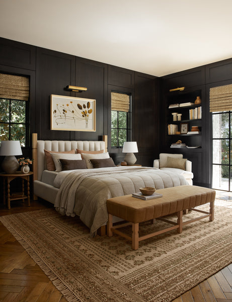 #size::cal-king #size::king #color::natural #size::queen | The Ambleside natural linen bed sits atop a patterned rug in a room with accented black wooden walls and a orange linen bench