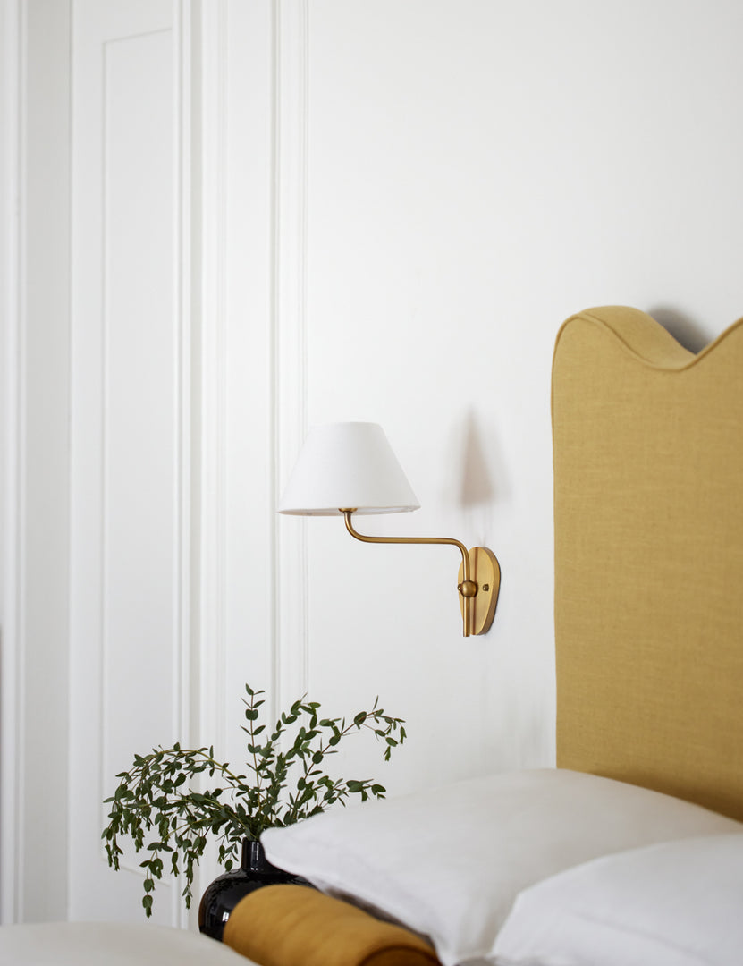 #color::white #finish::brass | The Magdalene brass single sconce is mounted to the right of a golden linen bed on a white wall