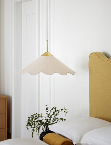 #color::brass #size::20-w | Panya pendant light hangs in a bedroom with white walls, a golden linen framed bed, and a house plant in a glossy black vase